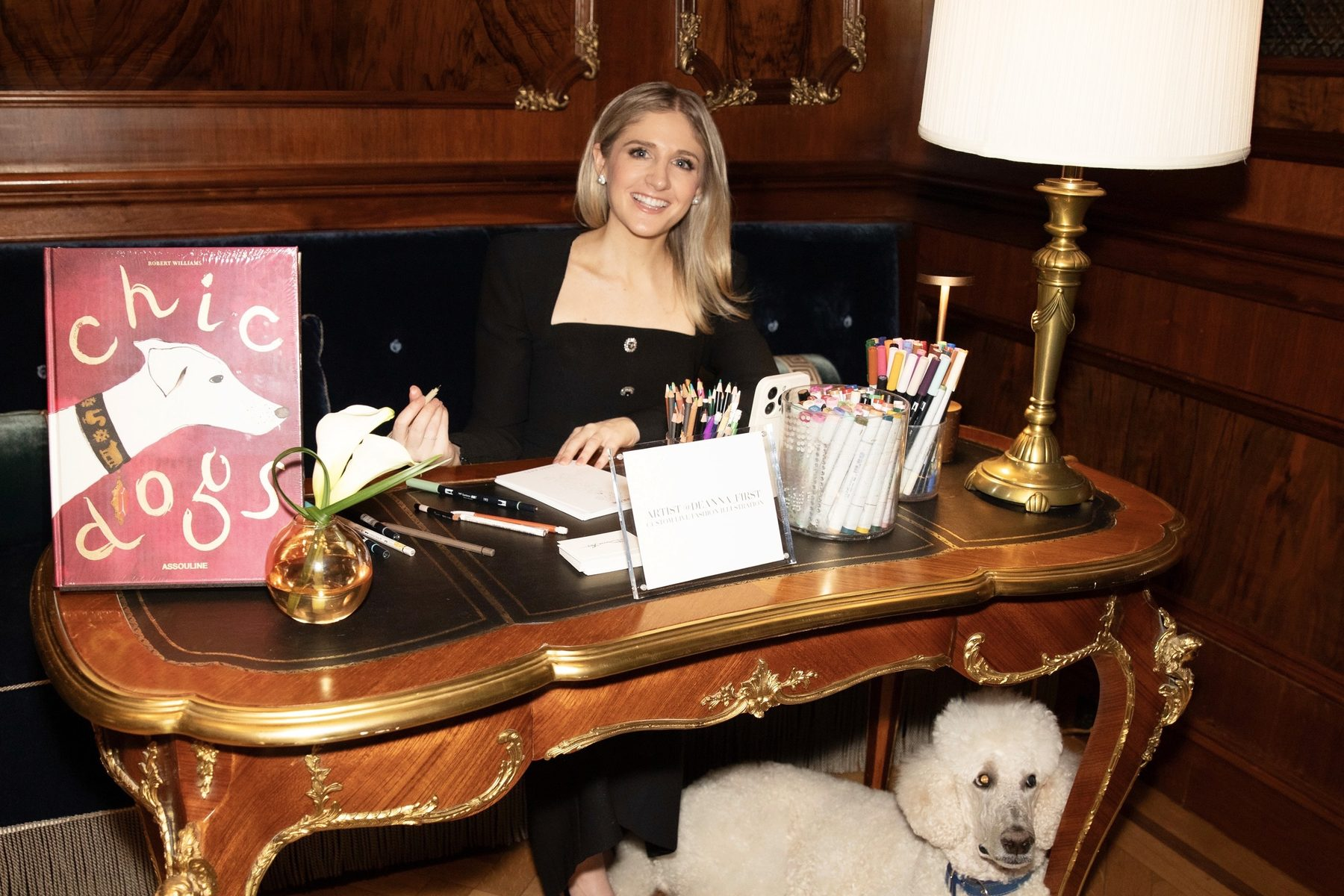 Fashion illustrator Deanna First at her desk with pens and markers, her poodle at her feet