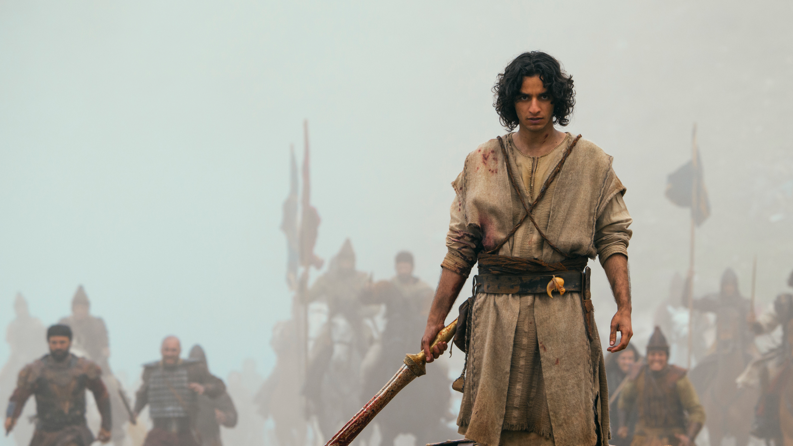 A young man with dark curly hair, his tunic stained with blood, holds a sword as warriors advance through the mist behind him