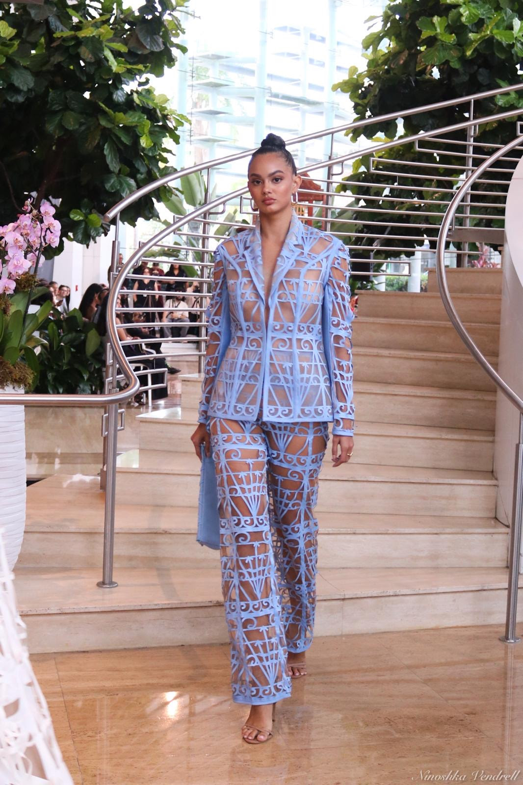 A model walks in a light blue laser-cut lattice blazer and wide-leg trousers in front of a curved marble staircase