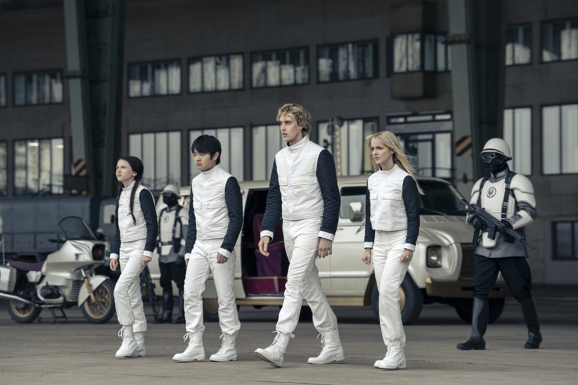 Four District 12 tributes in white uniforms with black sleeves walk across a tarmac, flanked by armored Peacekeepers.